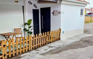 A building with a white exterior, two black doors, a wooden fence, a table, and a chair outside.