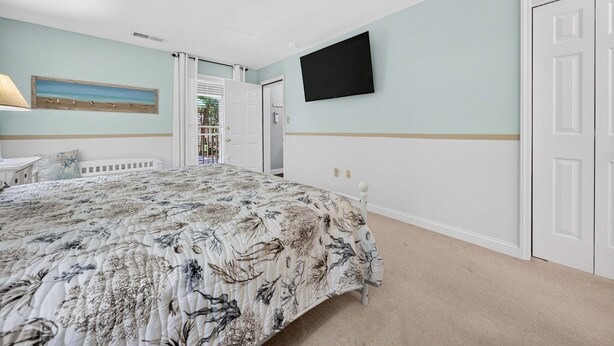 A bedroom with a bed, a TV mounted on the wall, and a door leading to a balcony. - 127-3 Puttin On The Ritz 2 Bedroom Condo (Pawleys Island)