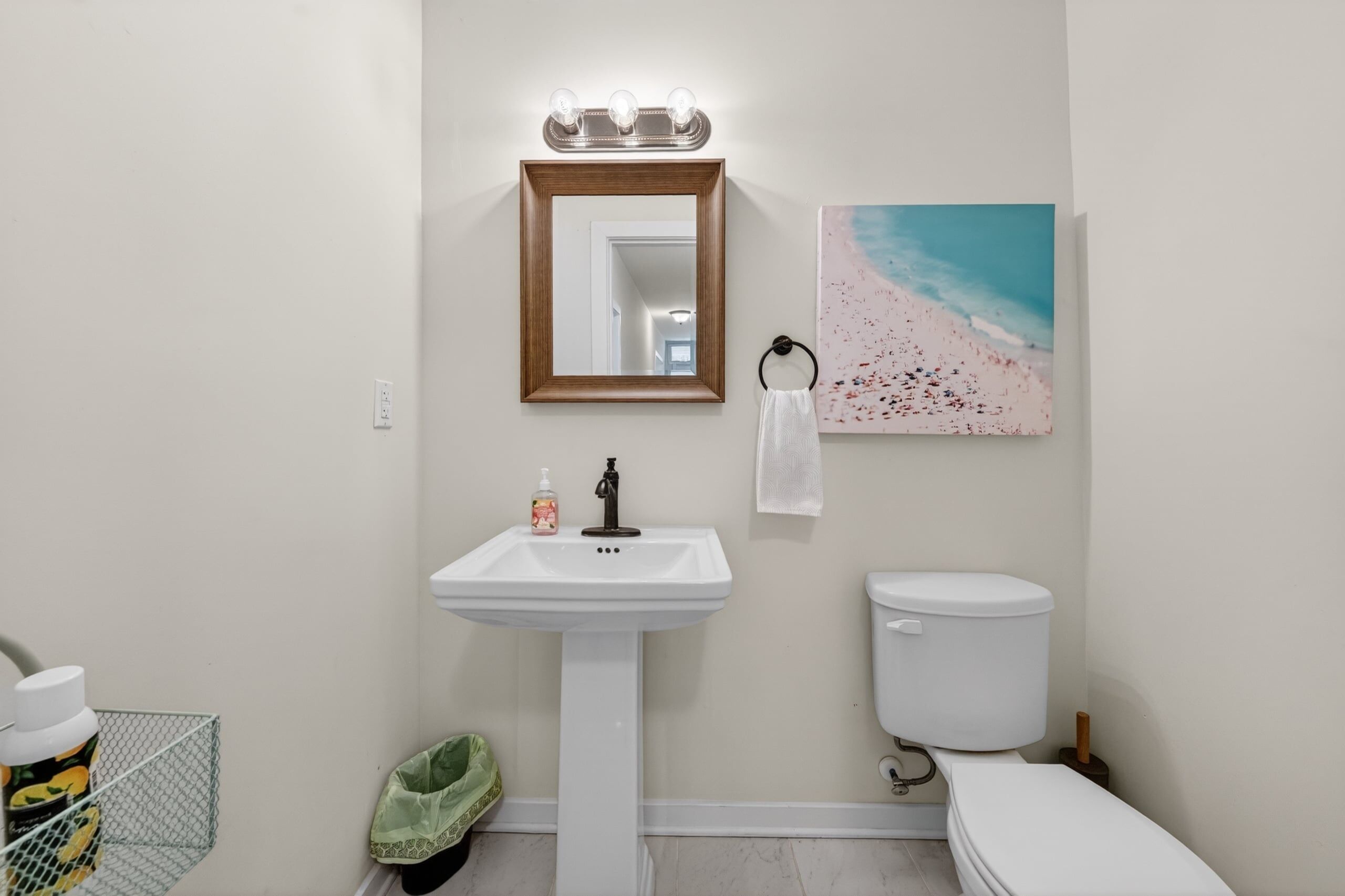 Un baño con un lavamanos blanco, un inodoro, un espejo con iluminación y un cuadro con motivos playeros.
