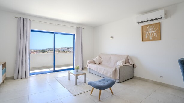 A modern living room with a sofa, a small table, a blue ottoman, and a large window with curtains. - #412 Apartment with Pool | 1BR | Near Oura & the Beaches (Albufeira)