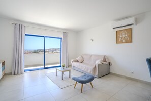 A modern living room with a sofa, a small table, a blue ottoman, and a large window with curtains. - #412 Apartment with Pool | 1BR | Near Oura & the Beaches (Albufeira)