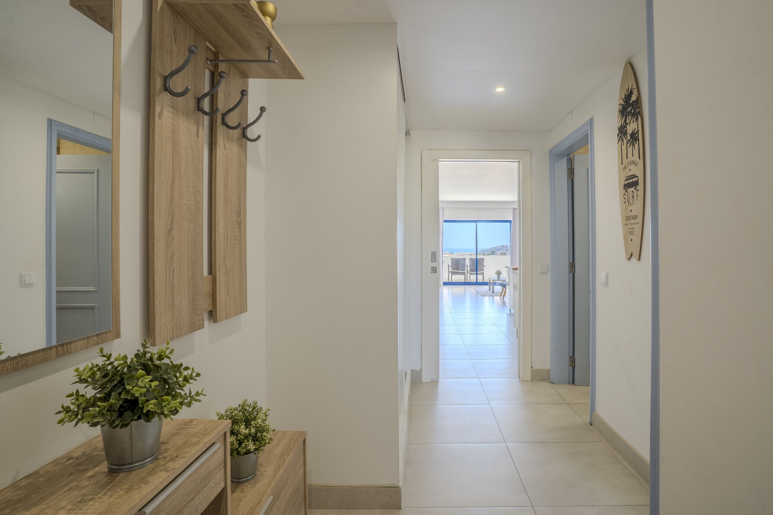 A hallway with a wooden bench, potted plants, a wall-mounted mirror, and a surfboard decoration.