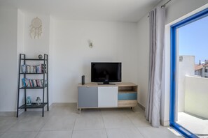 A modern living room with a flat-screen TV on a wooden cabinet, a bookshelf with books, a decorative wall hanging, and a view through a glass door. - #412 Apartment with Pool | 1BR | Near Oura & the Beaches (Albufeira)