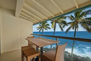 Ein Balkon mit Holztisch und Korbstühlen, mit Blick auf den Ozean und Palmen.
