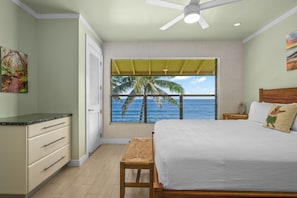 A bedroom with a bed, a desk, a chair, a window with a view of the ocean and palm trees, and a ceiling fan. - Top-Floor Oceanfront Condo with Panoramic Views & Heated Pool (Koloa)