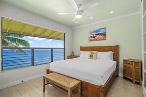 A bedroom with a large window overlooking the sea, a bed, a bench, a ceiling fan, and a nightstand. - Top-Floor Oceanfront Condo with Panoramic Views & Heated Pool (Koloa)