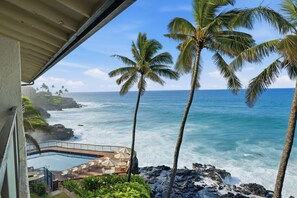 A balcony with a view of the ocean, palm trees, and a pool area with lounge chairs and umbrellas. - Top-Floor Oceanfront Condo with Panoramic Views & Heated Pool (Koloa)