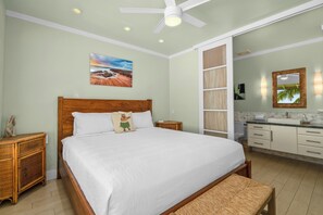 Ein Schlafzimmer mit einem großen Bett, einem hölzernen Kopfteil, einem Nachttisch, einer Ablage, einer Bank, einem Deckenventilator, einem Gemälde an der Wand und einem Badezimmer mit Waschbecken, Spiegel und Schränken.