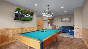 Une table de billard recouverte d’un drap vert, des meubles en bois, un téléviseur fixé au mur et un canapé bleu dans une pièce aux murs lambrissés.