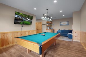 Ein Billardtisch mit grüner Spielbahn, Holzschränke, ein an der Wand befestigter Fernseher und ein blaues Sofa in einem Raum mit Holzvertäfelung.
