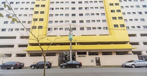 Um edifício com várias andares e fachada amarela, com um estacionamento subterrâneo e diversos carros estacionados na frente.