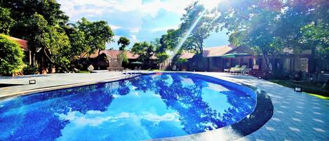 Una piscina con un deck de baldosas, rodeada de árboles y edificios.