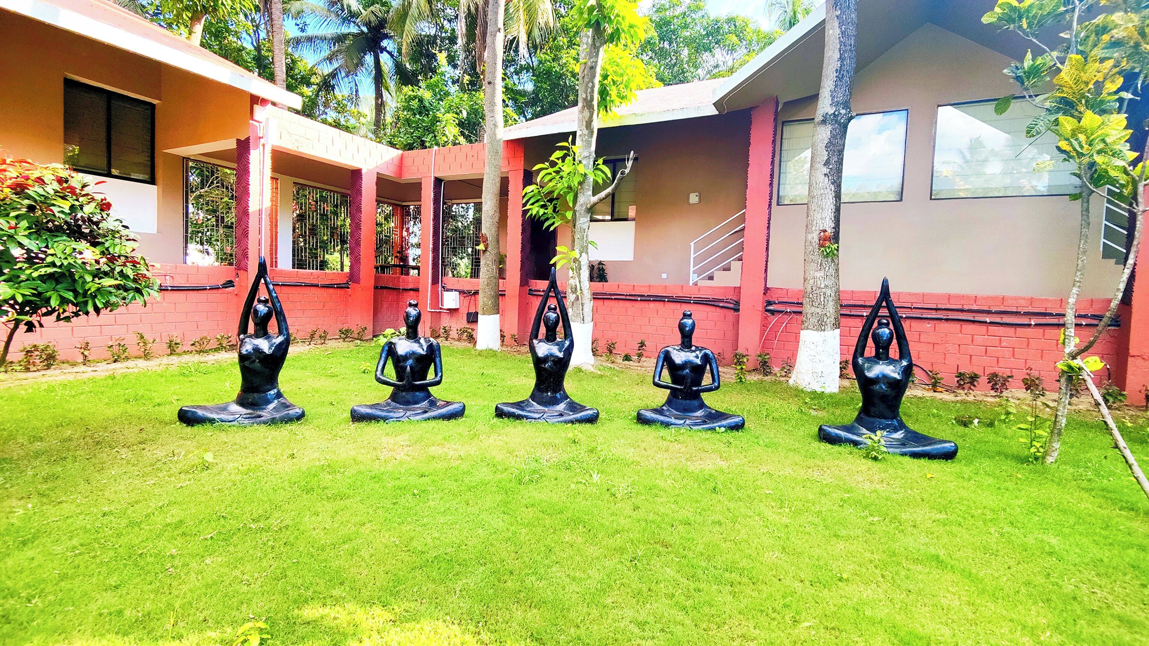 A courtyard with five black yoga statues on grass, surrounded by buildings with red and beige walls.