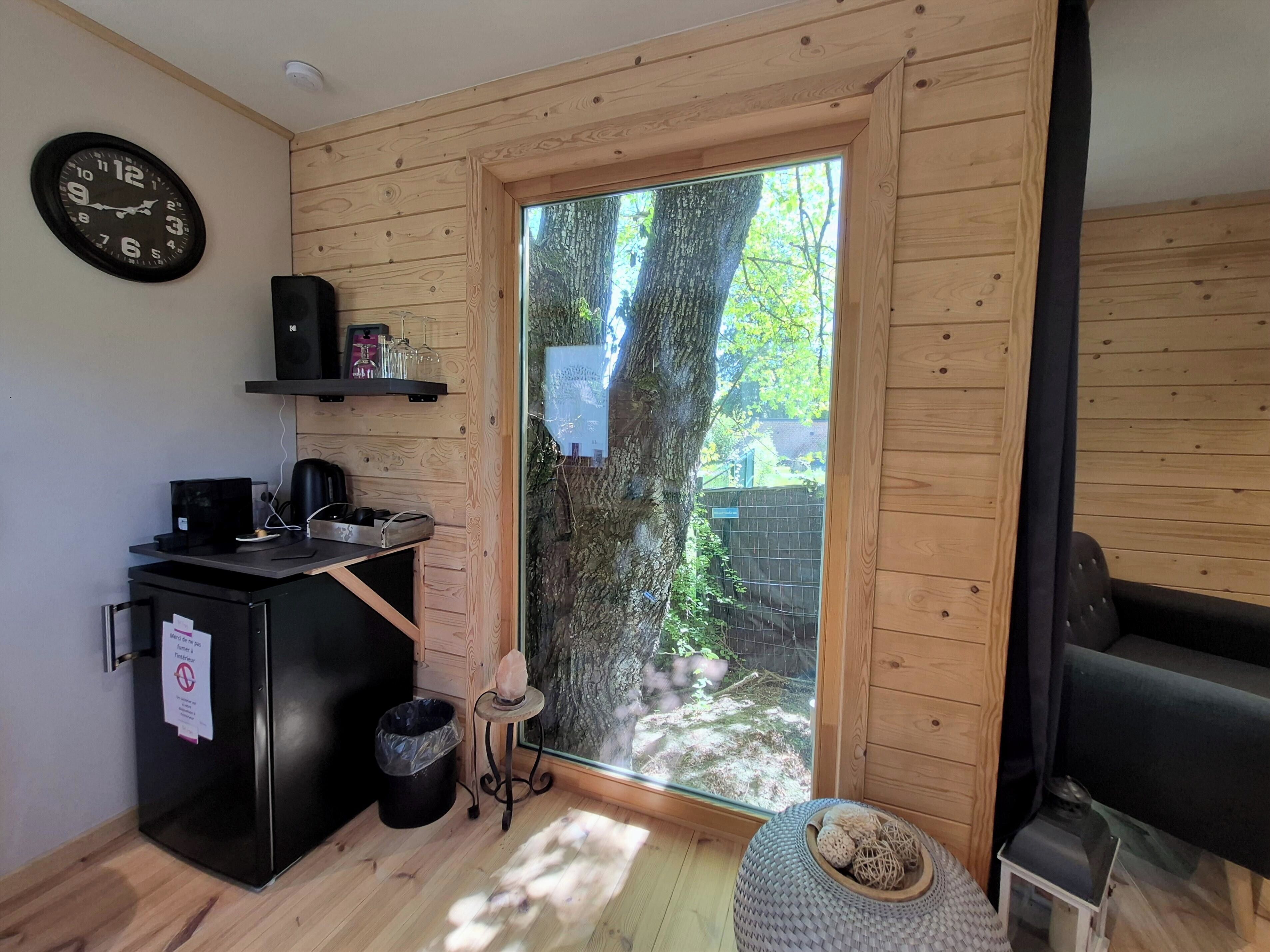 Une pièce de taille modeste, avec un intérieur en bois, un réfrigérateur noir, une machine à café, une horloge et une fenêtre donnant sur des arbres.