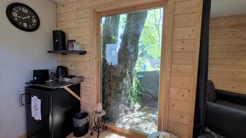 Une pièce de taille modeste, avec un intérieur en bois, un réfrigérateur noir, une machine à café, une horloge et une fenêtre donnant sur des arbres.