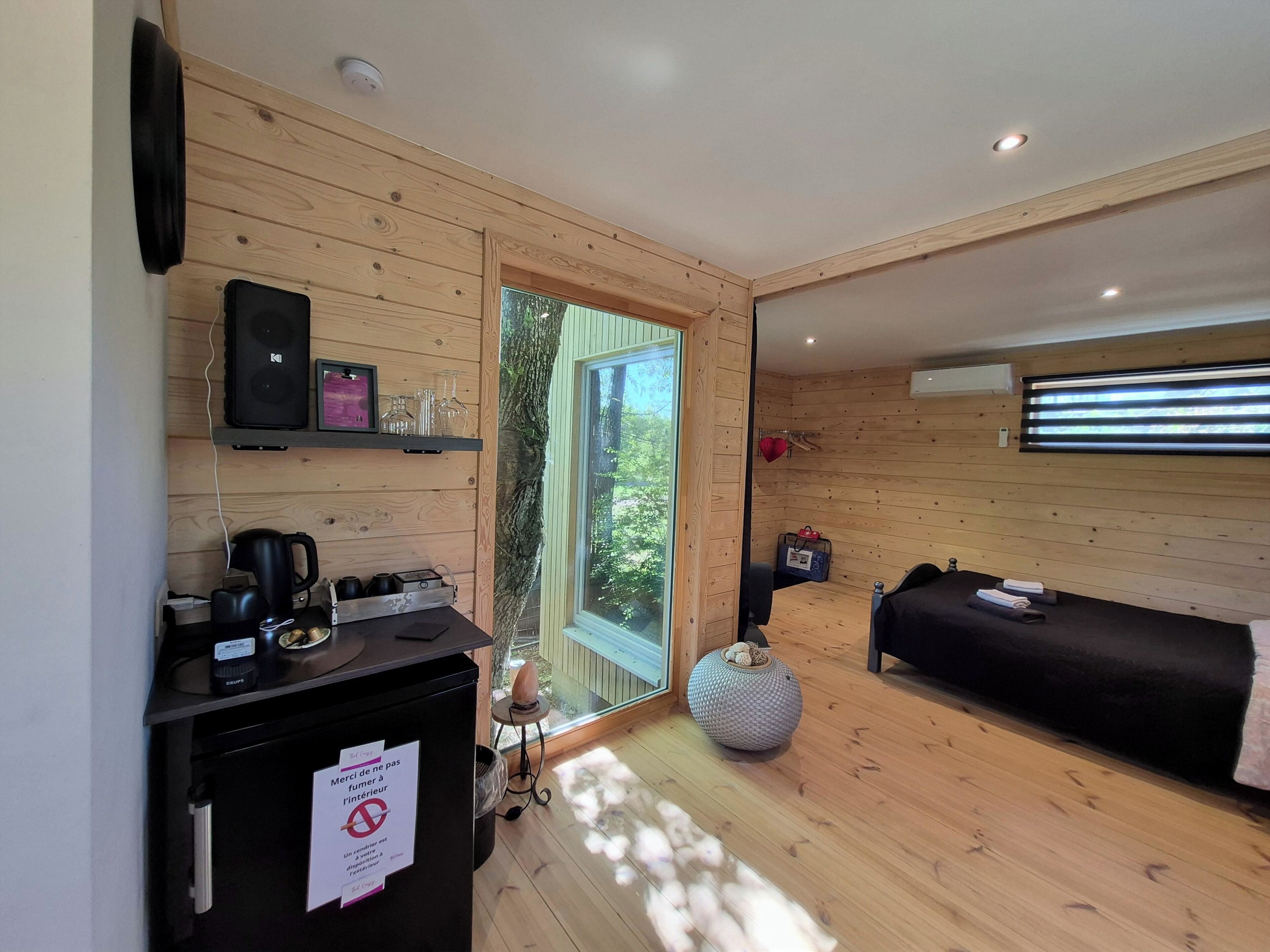 Una habitación moderna con detalles de madera, una cama, un escritorio con computadora y cafetera, y vistas a través de un ventanal.