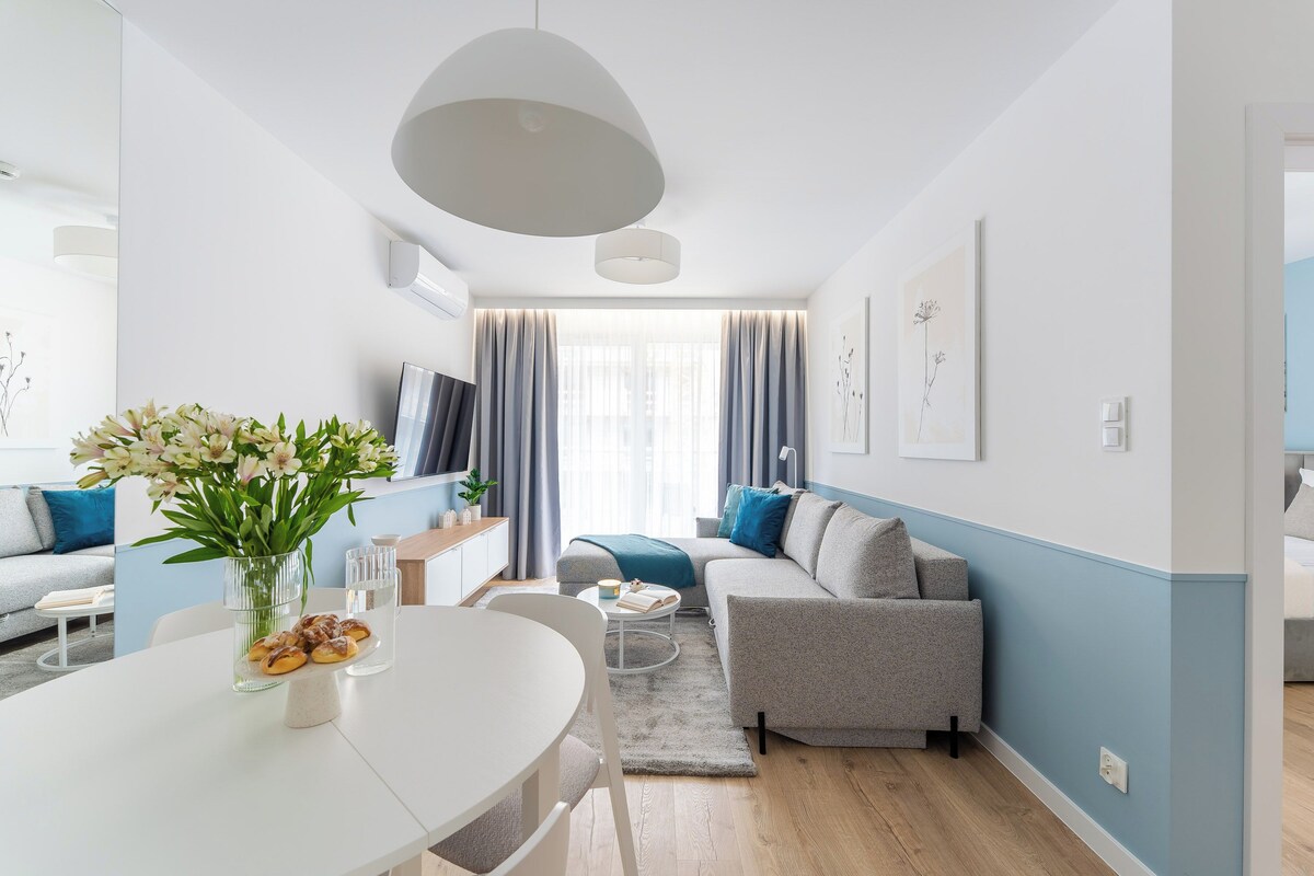 Ein modernes Wohnzimmer mit einer Couch, einem Esstisch mit Blumenvase und einem großen Fenster mit Vorhängen.