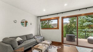 Un salon moderne avec un canapé gris, une table basse, un miroir rond et une grande fenêtre donnant sur un paysage verdoyant.