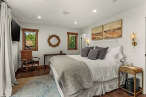 A bedroom with a large bed, a chair, a desk, and a view of greenery outside.