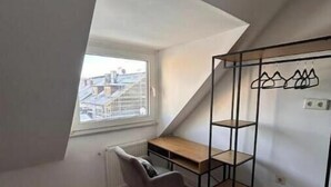 Ein kleines Zimmer im Dachgeschoss mit geneigter Decke, einem Holztisch, einem Stuhl und einem Metallregal.