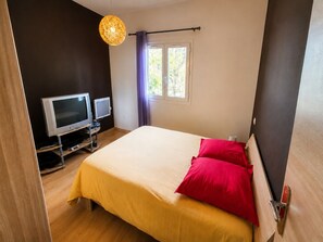 A bedroom with a bed, two televisions on a stand, a window with curtains, and a hanging light fixture. - Apartment, 2 Bedrooms for 3–4 Guests, Sauna & Ground-Floor Hot Tub (Le Tampon)