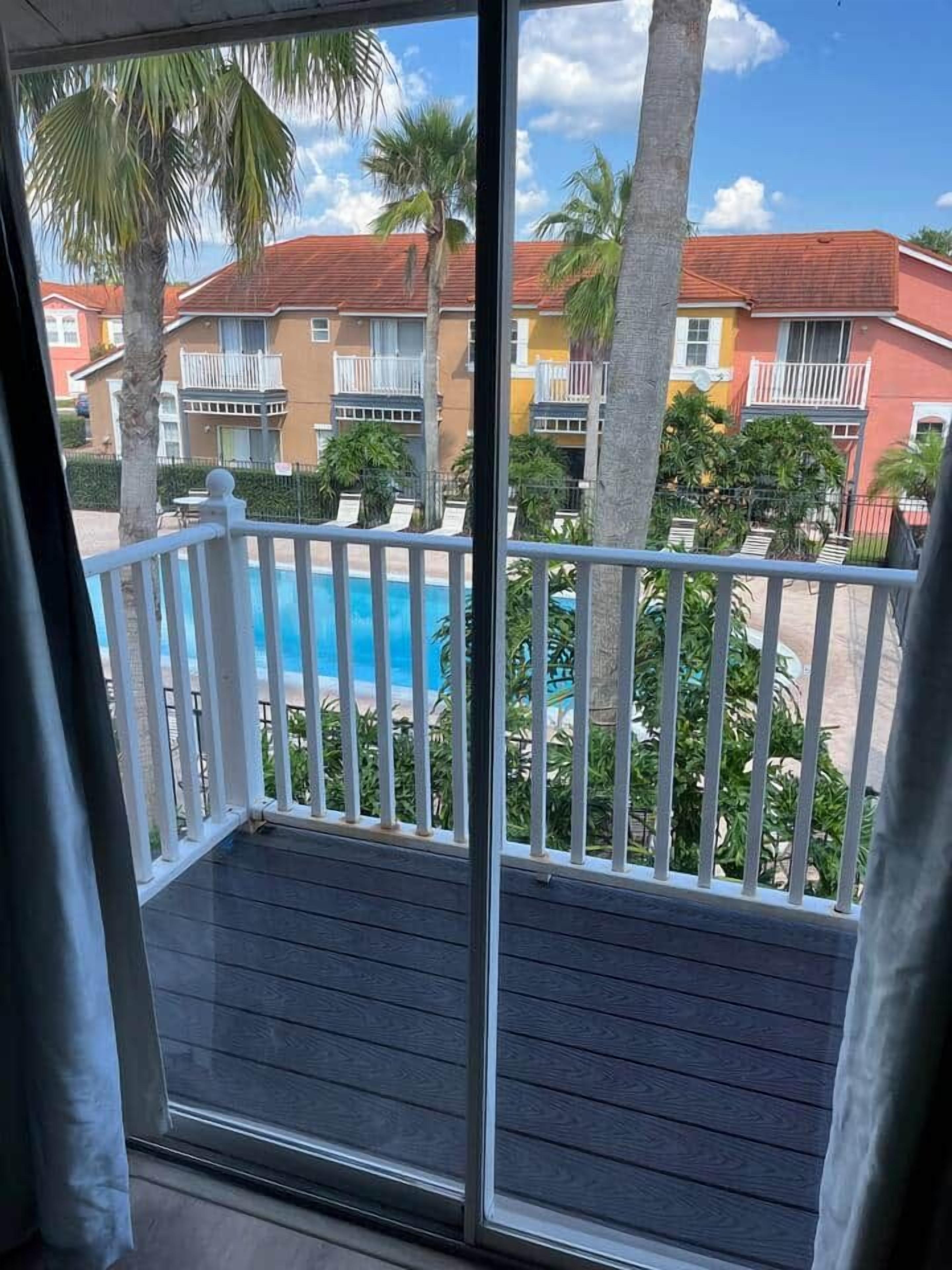Un balcon avec vue sur une piscine, des palmiers et des immeubles aux façades colorées.