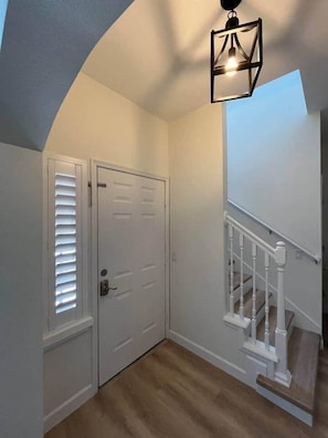 Un couloir avec une porte blanche, un escalier avec des rampes blanches et un luminaire suspendu.