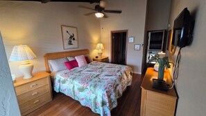 Een slaapkamer met een bed, nachtlampen, een plafondventilator, een televisie en een ingelijst kunstwerk aan de muur.