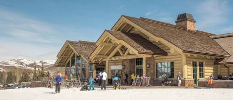 A lodge with a stone facade and wooden beams, featuring a large entrance and a ski slope in the foreground.