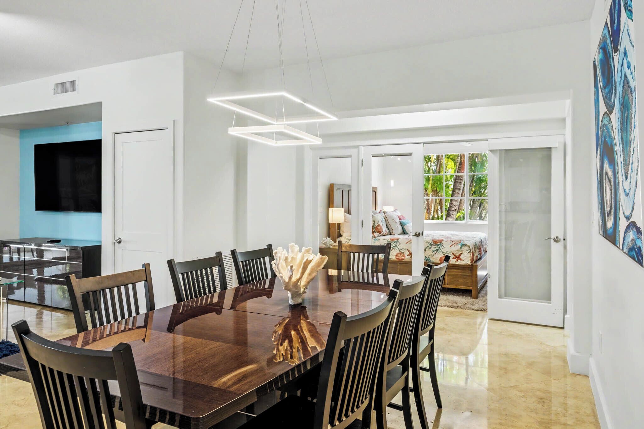 Un espace repas moderne avec une table et des chaises en bois, un élément décoratif central et une vue sur une chambre à coucher avec un lit et une fenêtre.