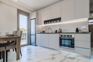 A modern kitchen with a built-in oven, a dining area with a table and chairs, and a view of the outdoors through a sliding door. - Ireon Luxury Suites 1 by RentalsPro - Nea Plagia Halkidiki (Nea Plagia)