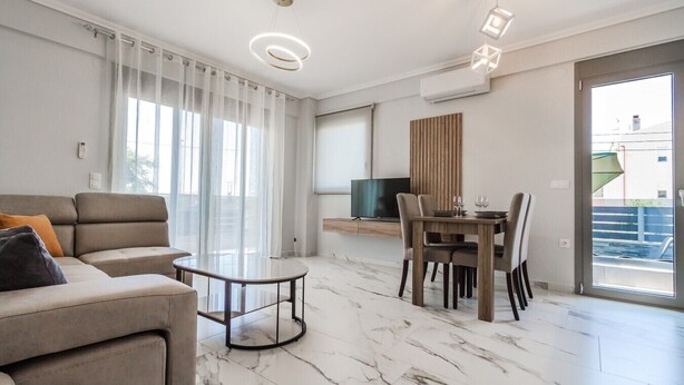 A modern living room with a sofa, a glass coffee table, a dining table with chairs, and a TV. - Ireon Luxury Suites 1 by RentalsPro - Nea Plagia Halkidiki (Nea Plagia)