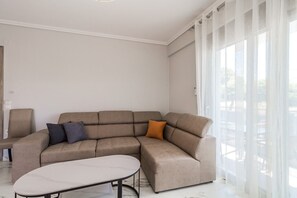 A modern living room with a sectional sofa, a small table, and sheer curtains. - Ireon Luxury Suites 1 by RentalsPro - Nea Plagia Halkidiki (Nea Plagia)