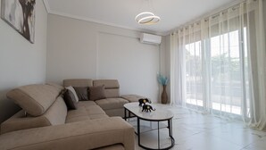A modern living room with a sectional sofa, a round coffee table, and a large window with sheer curtains. - Ireon Luxury Suites 2 by RentalsPro - Nea Plagia Halkidiki (Nea Plagia)