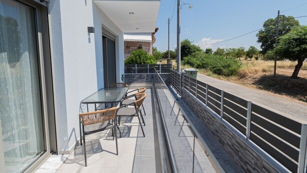A balcony with a table and chairs, a glass door, and a view of a road and greenery. - Ireon Luxury Suites 2 by RentalsPro - Nea Plagia Halkidiki (Nea Plagia)