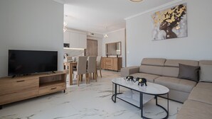 A modern living room with a flat-screen TV, a sofa, a coffee table, and a dining area with chairs and a wooden cabinet. - Ireon Luxury Suites 2 by RentalsPro - Nea Plagia Halkidiki (Nea Plagia)