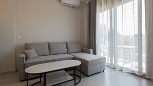 A modern living room with a grey sofa, a small table, and a large window with sheer curtains. - Ireon Luxury Suites 4 by RentalsPro - Nea Plagia Halkidiki (Nea Plagia)