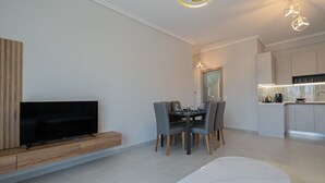 A modern living room with a flat-screen TV, a dining area with a table set for four, and a kitchen with a countertop and cabinets. - Ireon Luxury Suites 4 by RentalsPro - Nea Plagia Halkidiki (Nea Plagia)