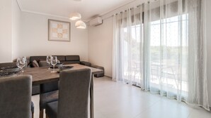 A dining area with a table set for four, a sofa, and a large window with sheer curtains. - Ireon Luxury Suites 3 by RentalsPro - Nea Plagia Halkidiki (Nea Plagia)