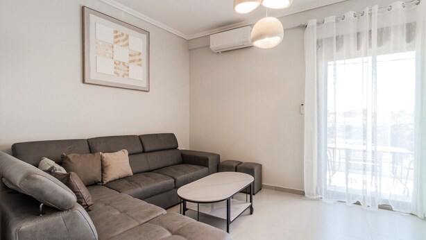 A modern living room with a grey sectional sofa, a small round table, and a large window with sheer curtains. - Ireon Luxury Suites 3 by RentalsPro - Nea Plagia Halkidiki (Nea Plagia)
