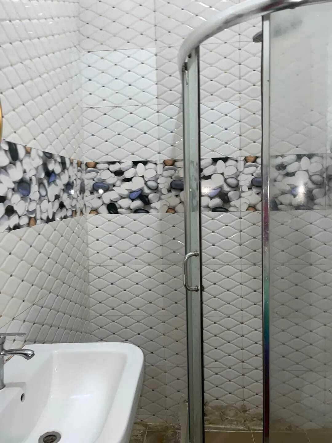 Une salle de bain avec un lavabo blanc, une cabine de douche en verre et un mur carrelé orné d’un motif de galets.