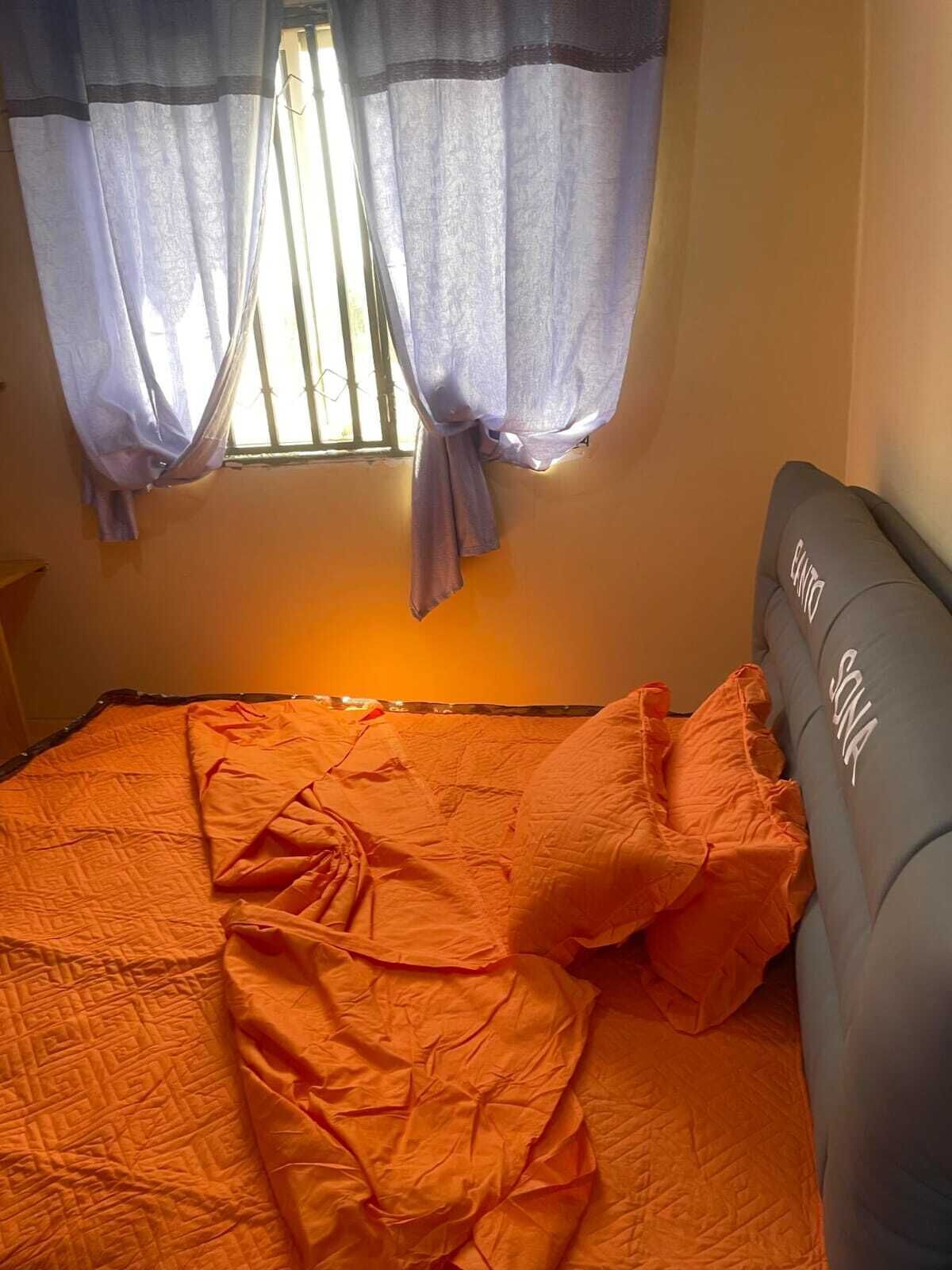 Una cama con colcha y almohadas naranjas, cabecera gris con la palabra 'SUNA' y una ventana con cortinas moradas.