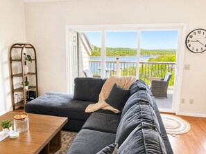 Ein Wohnzimmer mit einer Eckcouch, einem Holztisch, einem Bücherregal und Blick auf einen See durch eine Glastür.