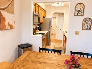 Une cuisine avec des meubles de rangement en bois, un réfrigérateur et un four à micro-ondes. On y trouve une table à manger avec des chaises et une poubelle.