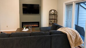 A living room with a sectional sofa, a flat-screen TV, a fireplace, and a bookshelf. - Charming 4BR Lakefront Condo Getaway- Near Boat Marina and Silver Dollar City. (Branson)