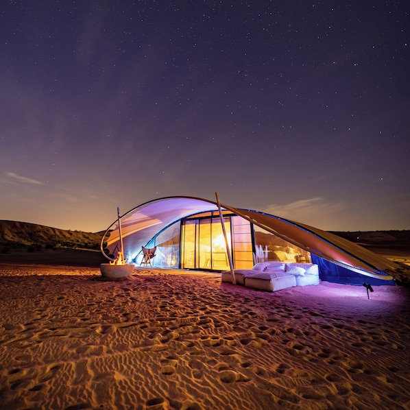 A dome-shaped tent with a glass entrance, a fire pit, and a bed inside.