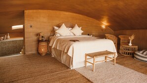 Un dormitorio con techo abovedado, una cama con almohadas, una mesita de noche, una silla de mimbre y una pequeña mesa redonda con un florero.