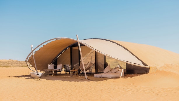Tienda de campaña en el desierto con entrada arqueada, un área de comedor con sillas y una mesa, y un cuenco grande.