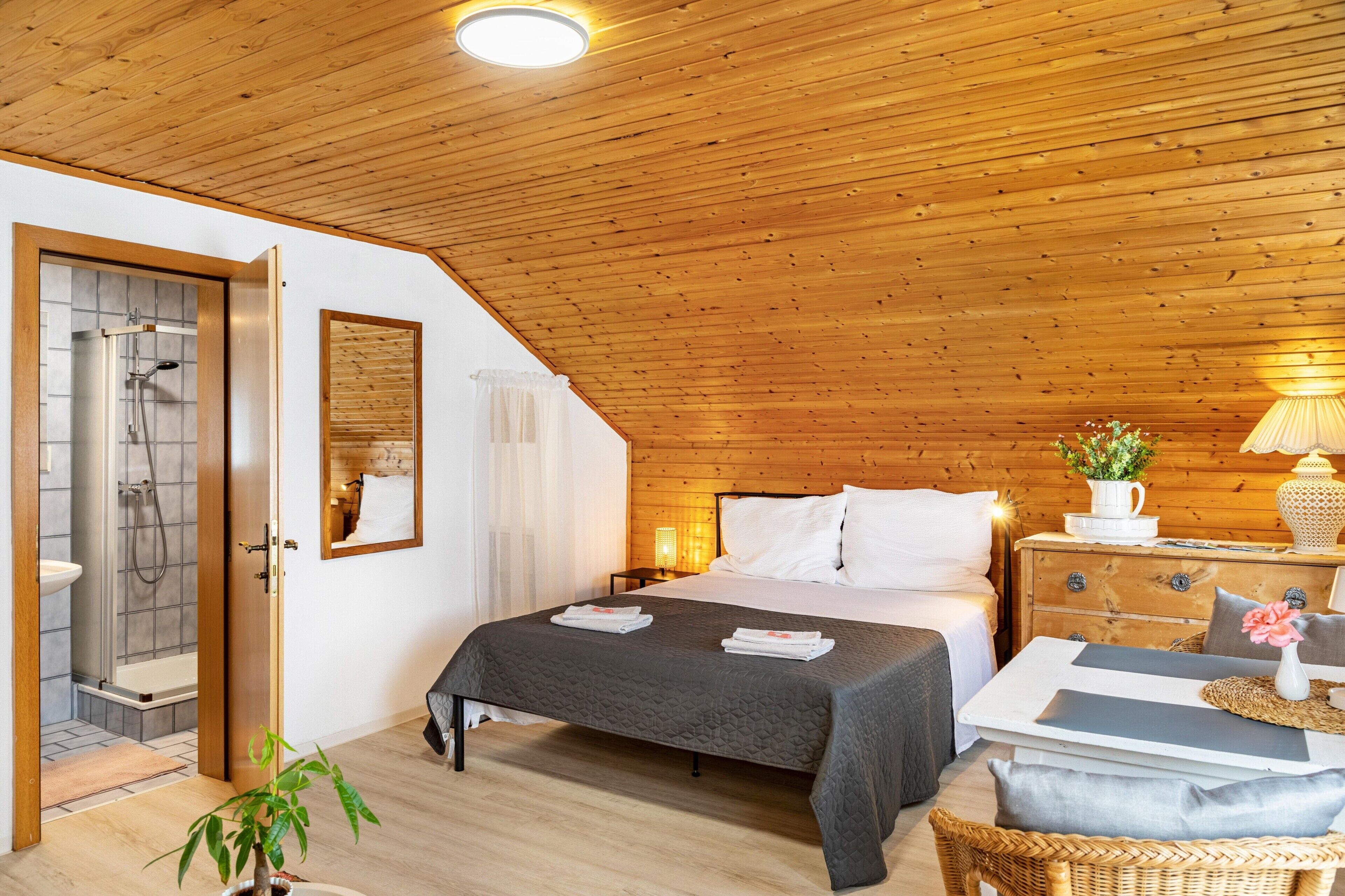 Et hyggeligt soveværelse med træloft, en seng med hvide sengetøj, et natbord med lampe og udsigt til et badeværelse med bruser.