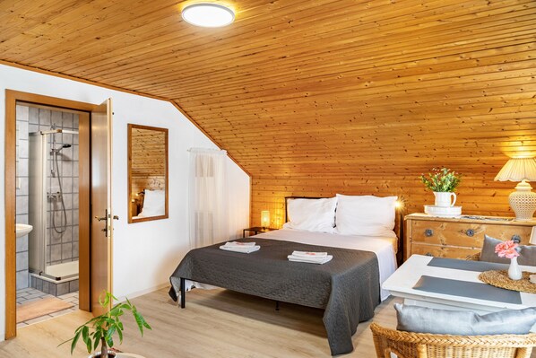 A cozy bedroom with a wooden ceiling, a bed with white linens, a bedside table with a lamp, and a view of a bathroom with a shower.